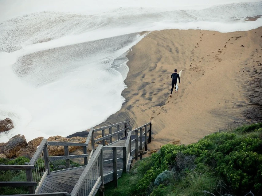 A lone surfer walks along a sandy beach toward the ocean, carrying a surfboard. White waves roll in, meeting the shore, with greenery visible.