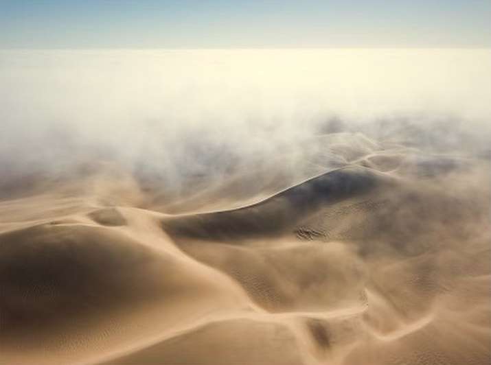 Dramatic aerial view of sand dunes with low-lying fog in a desert landscape, highlighting the texture and scale of the natural environment.
