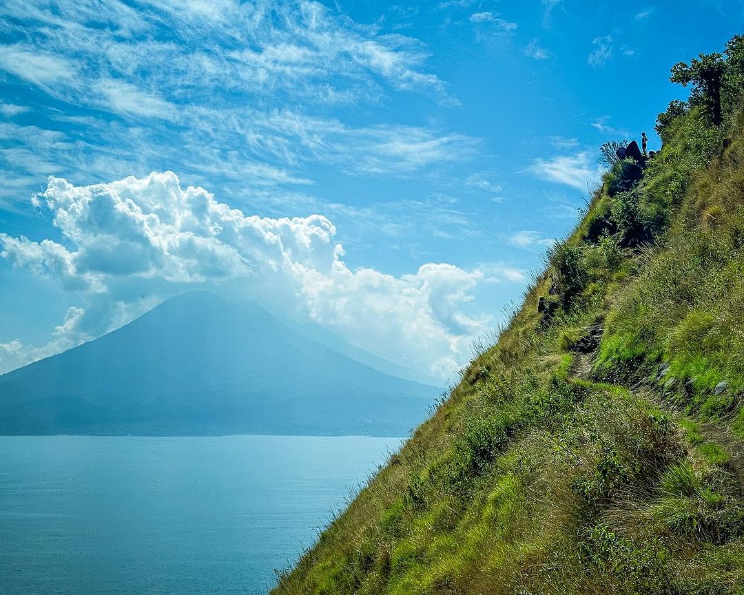 A high-altitude hiking trail on a steep grassy ridge overlooking a large lake and a distant volcanic mountain under a bright blue sky.