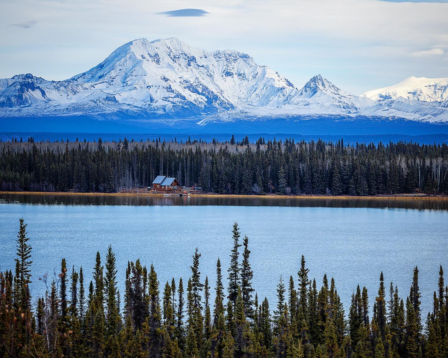 Scenic landscape featuring a tranquil lake surrounded by a dense forest and snow-capped mountains in the background under a bright sky.