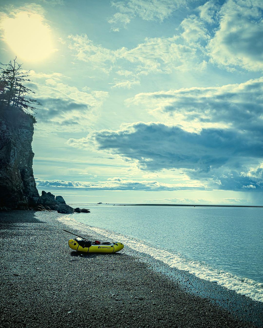 Yellow packraft sits on a rocky beach next to the ocean under a cloudy sky with a rocky cliff in the background on the left.