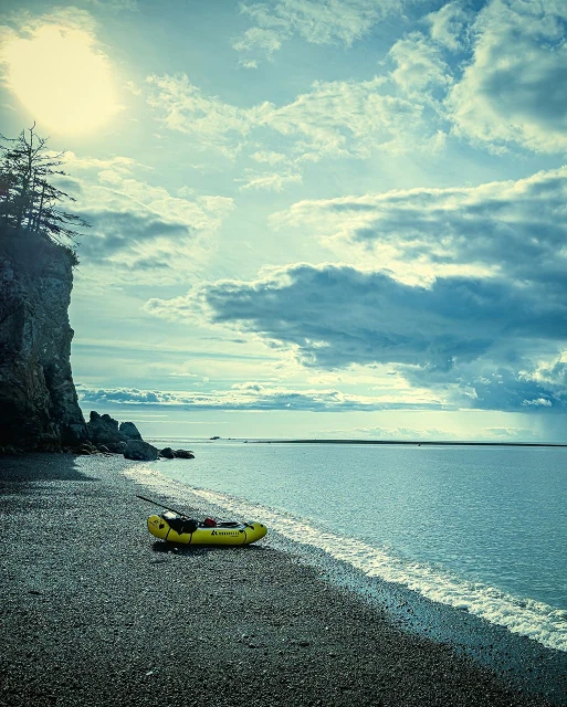 Yellow packraft sits on a rocky beach next to the ocean under a cloudy sky with a rocky cliff in the background on the left.