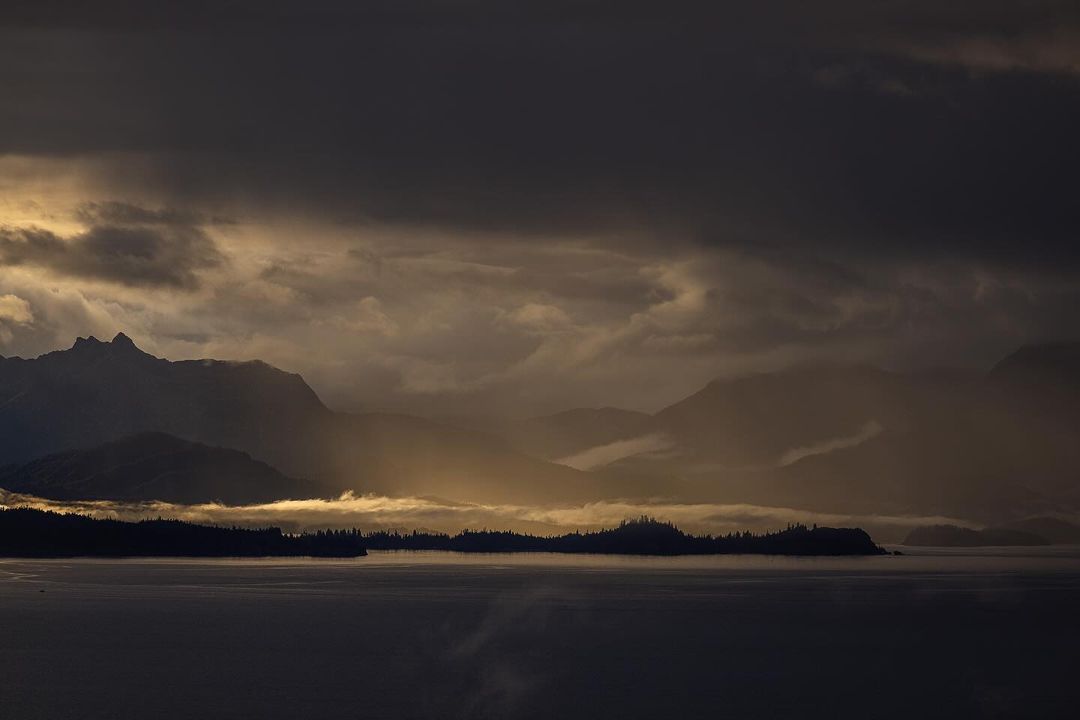 Dramatic landscape featuring silhouetted mountains and islands under a cloudy, moody sky with sunlight breaking through the clouds.