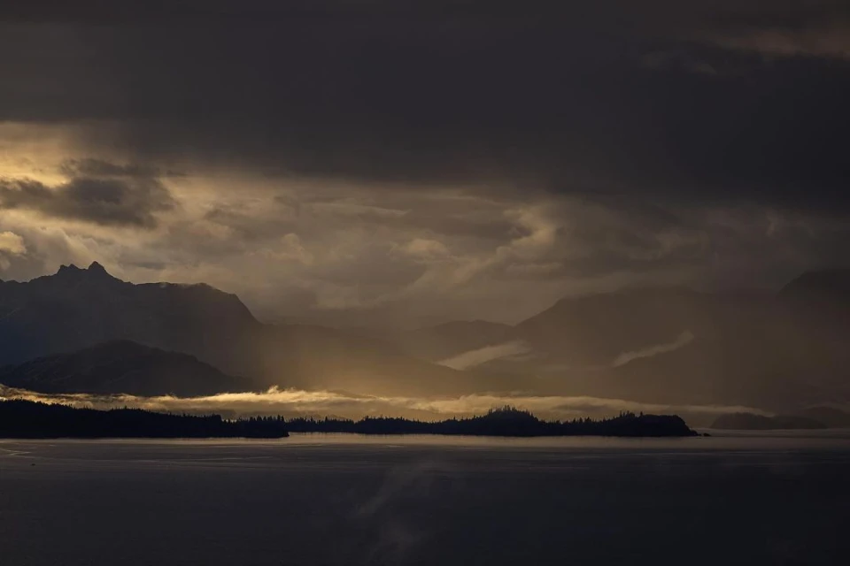 Dramatic landscape featuring silhouetted mountains and islands under a cloudy, moody sky with sunlight breaking through the clouds.