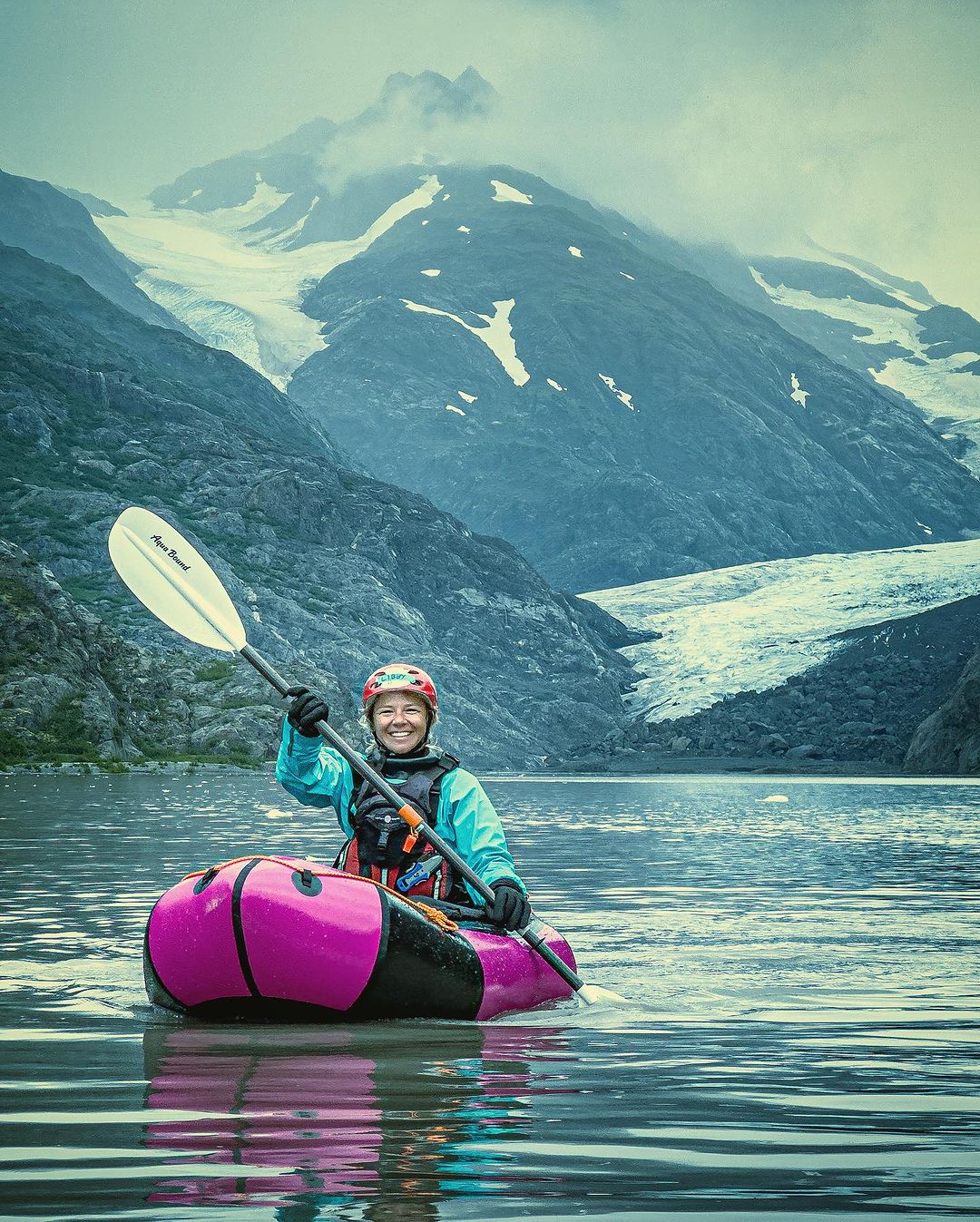 A woman smiles while paddling an inflatable kayak on a serene lake, set against a backdrop of majestic glacier-covered mountains and cloudy skies.