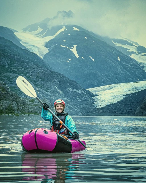 A woman smiles while paddling an inflatable kayak on a serene lake, set against a backdrop of majestic glacier-covered mountains and cloudy skies.