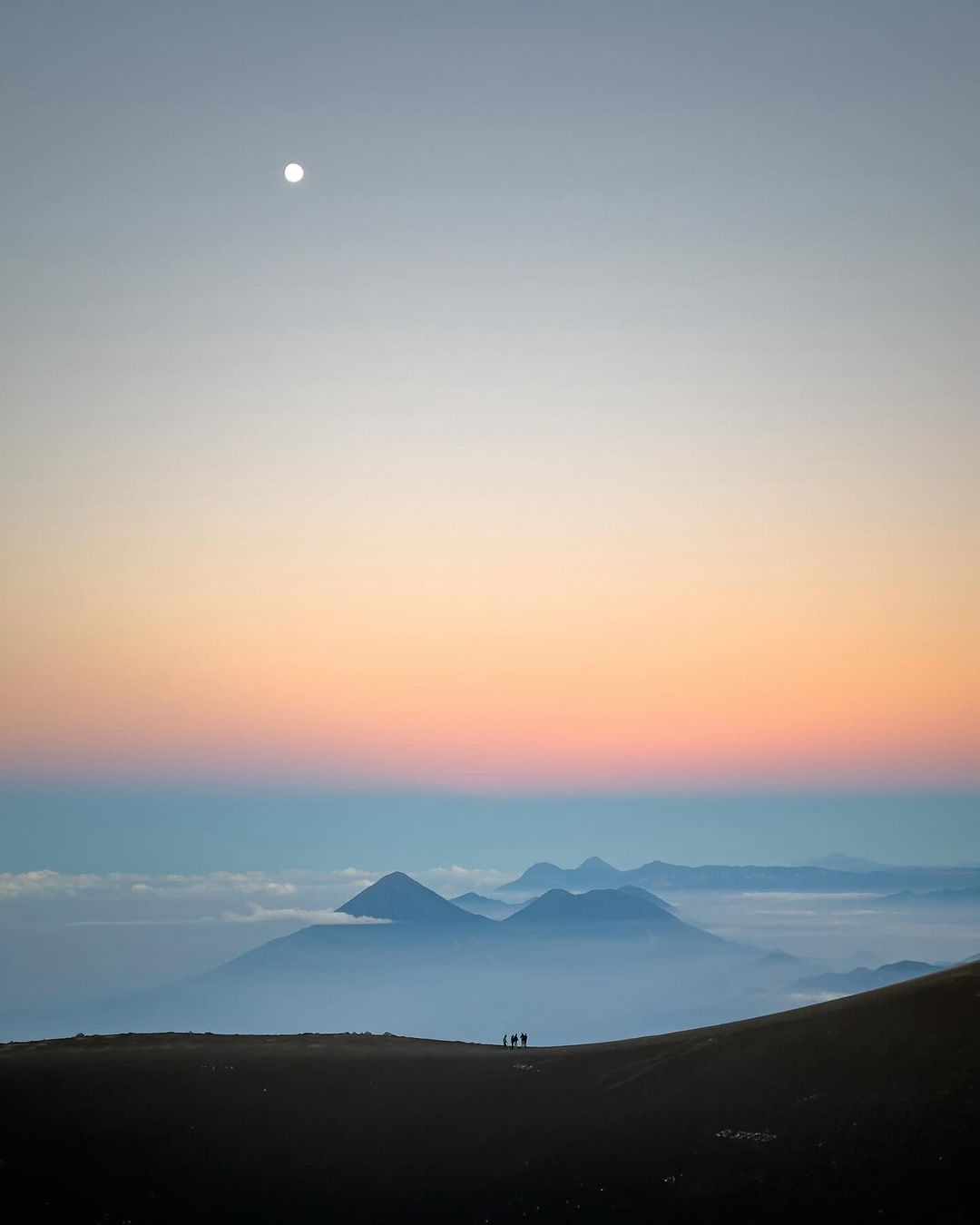 Silhouetted group of hikers on a ridge at sunset overlook a valley filled with low-lying clouds and distant mountains.