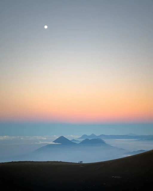 Silhouetted group of hikers on a ridge at sunset overlook a valley filled with low-lying clouds and distant mountains.