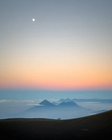 Silhouetted group of hikers on a ridge at sunset overlook a valley filled with low-lying clouds and distant mountains.