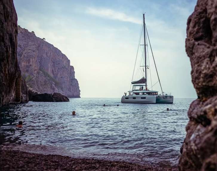 Scenic view of a sailboat anchored offshore with people swimming, near rocky cliffs and a pebble beach under a partly cloudy sky.