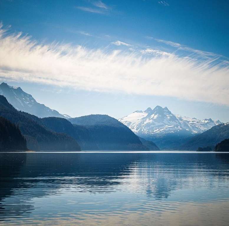 Scenic view of a calm lake surrounded by forested hills and snow-capped mountains under a blue sky with scattered clouds.