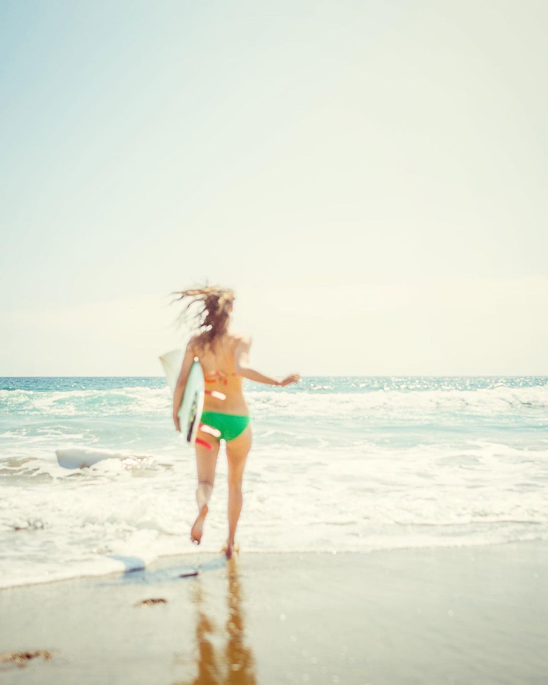 A woman runs into the ocean with a surfboard on a sunny day. The beach and waves frame the scene.