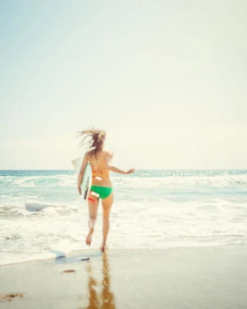 A woman runs into the ocean with a surfboard on a sunny day. The beach and waves frame the scene.