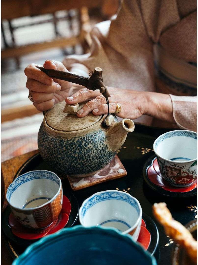 Adult human pours tea from ceramic kettle into traditional tea cups arranged on a dark serving tray in an indoor setting.