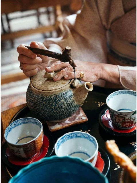 Adult human pours tea from ceramic kettle into traditional tea cups arranged on a dark serving tray in an indoor setting.