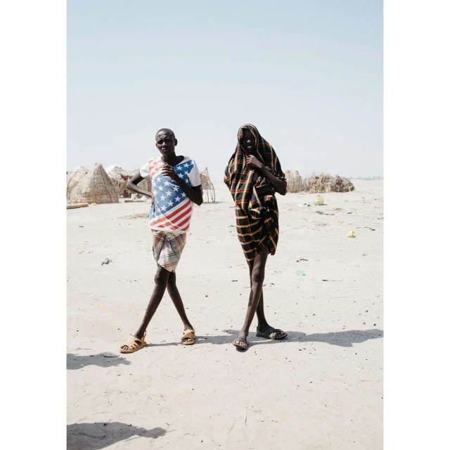 Two slender figures pose outdoors in an arid landscape. One wears a U.S. flag shirt, while the other is draped in a striped shawl.