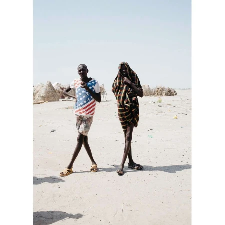 Two slender figures pose outdoors in an arid landscape. One wears a U.S. flag shirt, while the other is draped in a striped shawl.