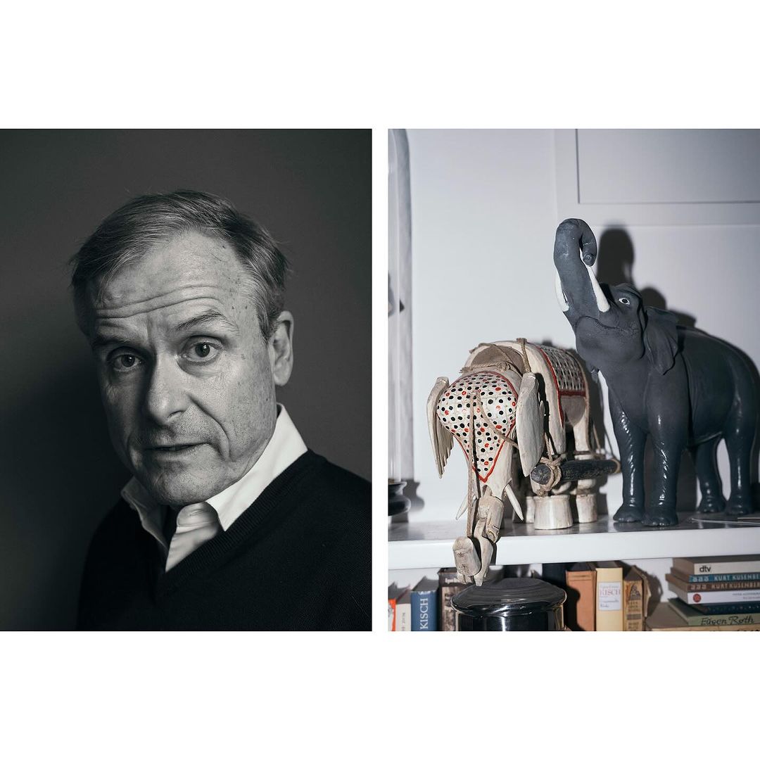 Monochrome portrait of an adult male with a neutral expression alongside a pair of decorative elephant figurines on a shelf indoors.