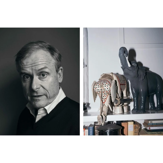 Monochrome portrait of an adult male with a neutral expression alongside a pair of decorative elephant figurines on a shelf indoors.