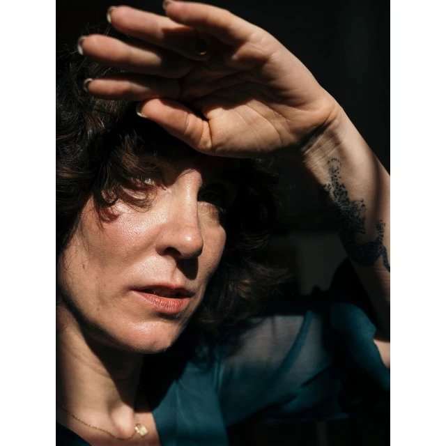 Close-up portrait of a woman with curly hair shielding her eyes from bright sunlight with her hand, casting shadows across her face.