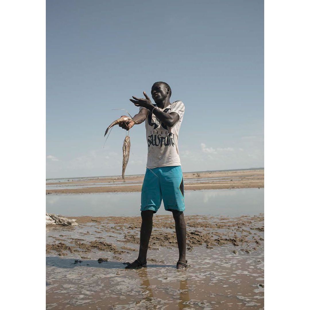 Adult male poses on a sandy beach holding two fish in his hand on a sunny day with the blue sky and water in the background.
