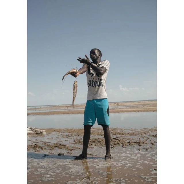 Adult male poses on a sandy beach holding two fish in his hand on a sunny day with the blue sky and water in the background.