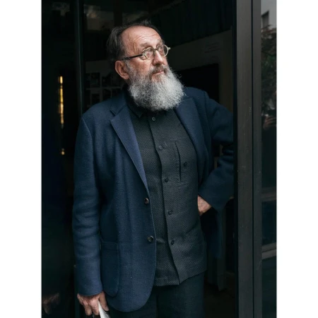 Elderly man with a long white beard wearing round glasses and a dark blue blazer standing in a doorway