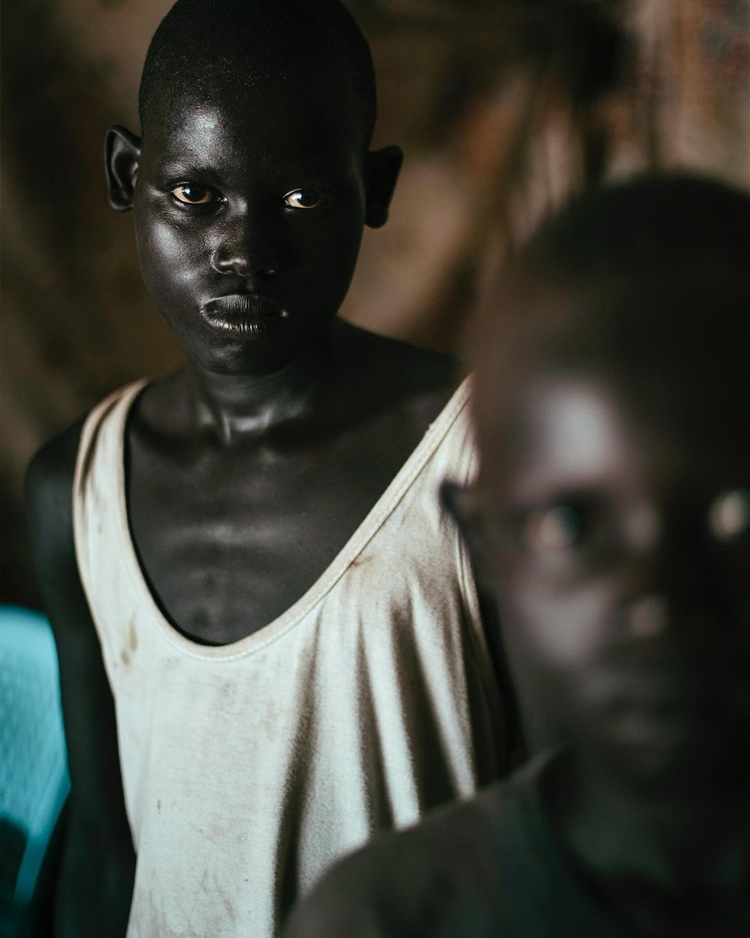 Striking portrait of two young Black children indoors, one in focus wearing a white tank top, with soft lighting and a blurred background.