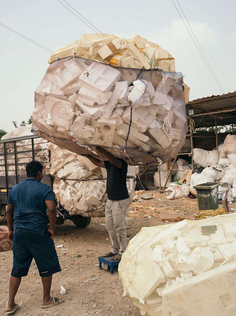 Two men in an outdoor setting handling a large bag filled with styrofoam pieces near a truck and building, with overcast sky overhead.