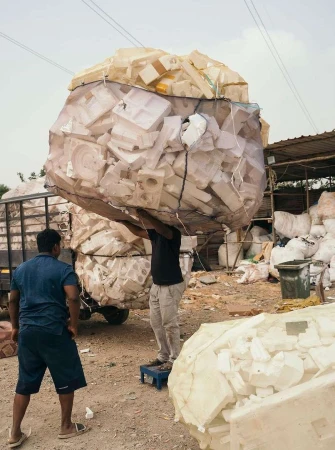 Two men in an outdoor setting handling a large bag filled with styrofoam pieces near a truck and building, with overcast sky overhead.