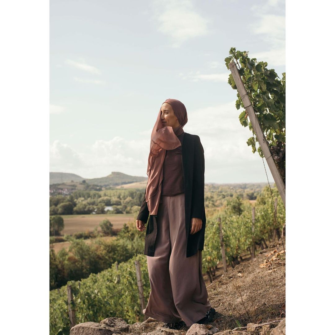 Adult woman wearing a headscarf, black jacket and wide-legged trousers standing outdoors near a vineyard and a distant landscape.