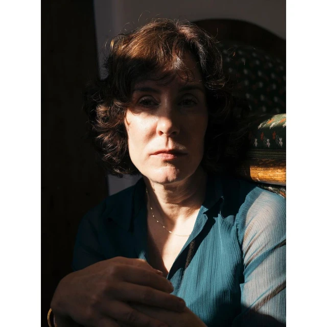 Portrait of a woman with dark hair and a teal top sitting in a chair indoors, illuminated by a beam of natural light.