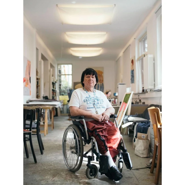 An adult in a wheelchair poses in an art studio, surrounded by tables, art supplies, and natural light from large windows.