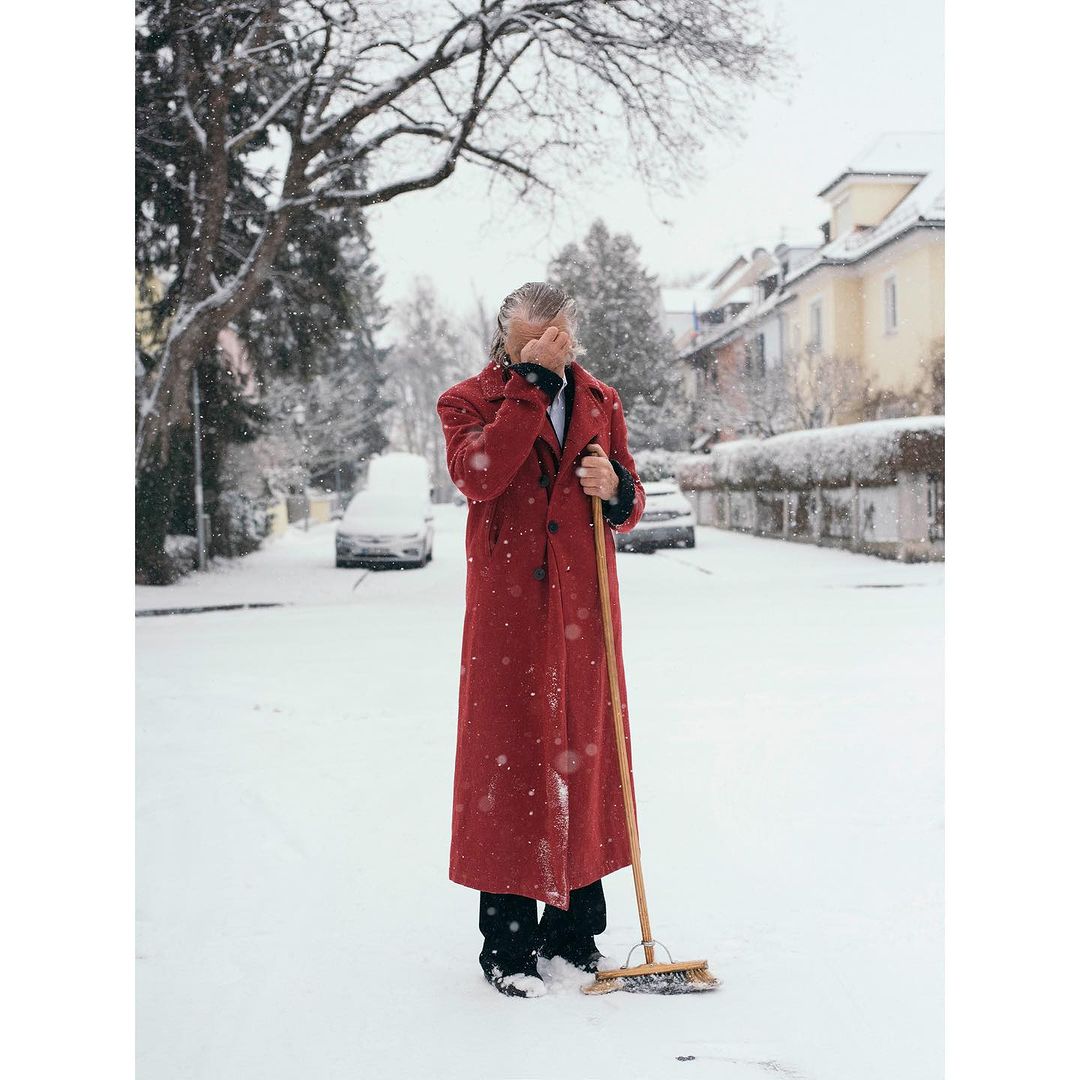 Man in a red coat sweeps snow on a tree-lined street with cars and houses in the background on a winter day with overcast lighting.