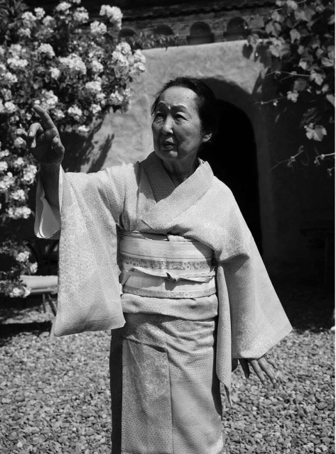 Monochrome portrait of a woman in a kimono standing outside a building with plants. Woman has her arms extended with an open expression.