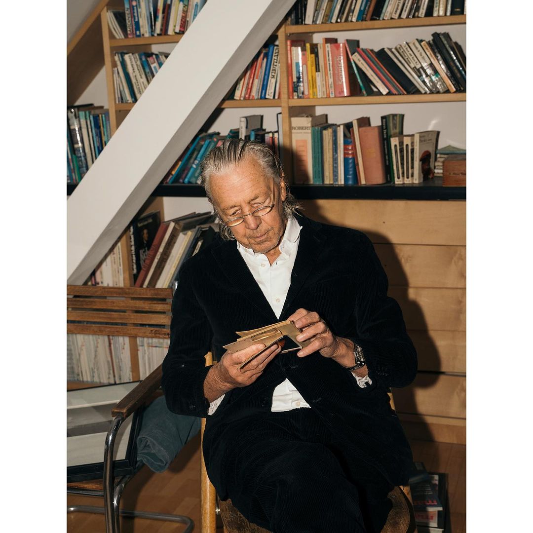 Older man with glasses sits indoors, examining cards, wearing a black velvet jacket with a bookshelf filled with books in background.