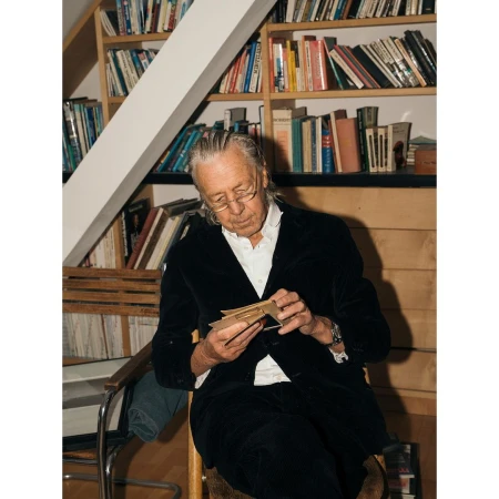 Older man with glasses sits indoors, examining cards, wearing a black velvet jacket with a bookshelf filled with books in background.