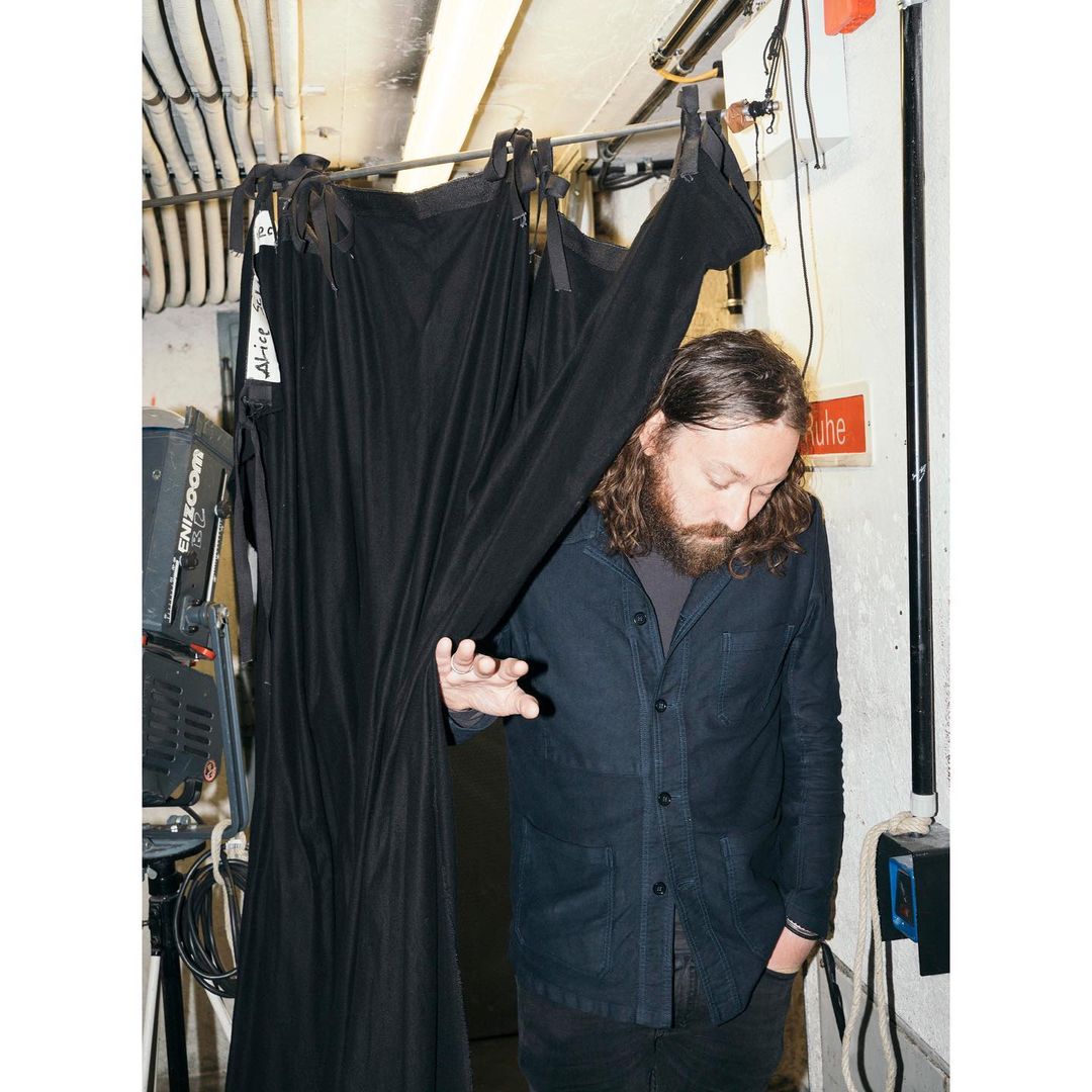 Man with a beard stands in front of a black curtain, with coiled cables and lighting equipment visible in the background of an indoor set.