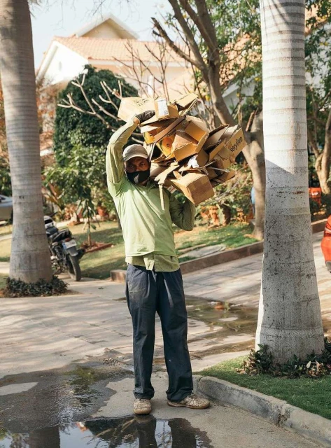 Man in a light green shirt carries a load of cardboard boxes on his shoulder, walking on a paved road with trees and buildings nearby.