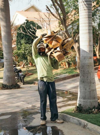Man in a light green shirt carries a load of cardboard boxes on his shoulder, walking on a paved road with trees and buildings nearby.
