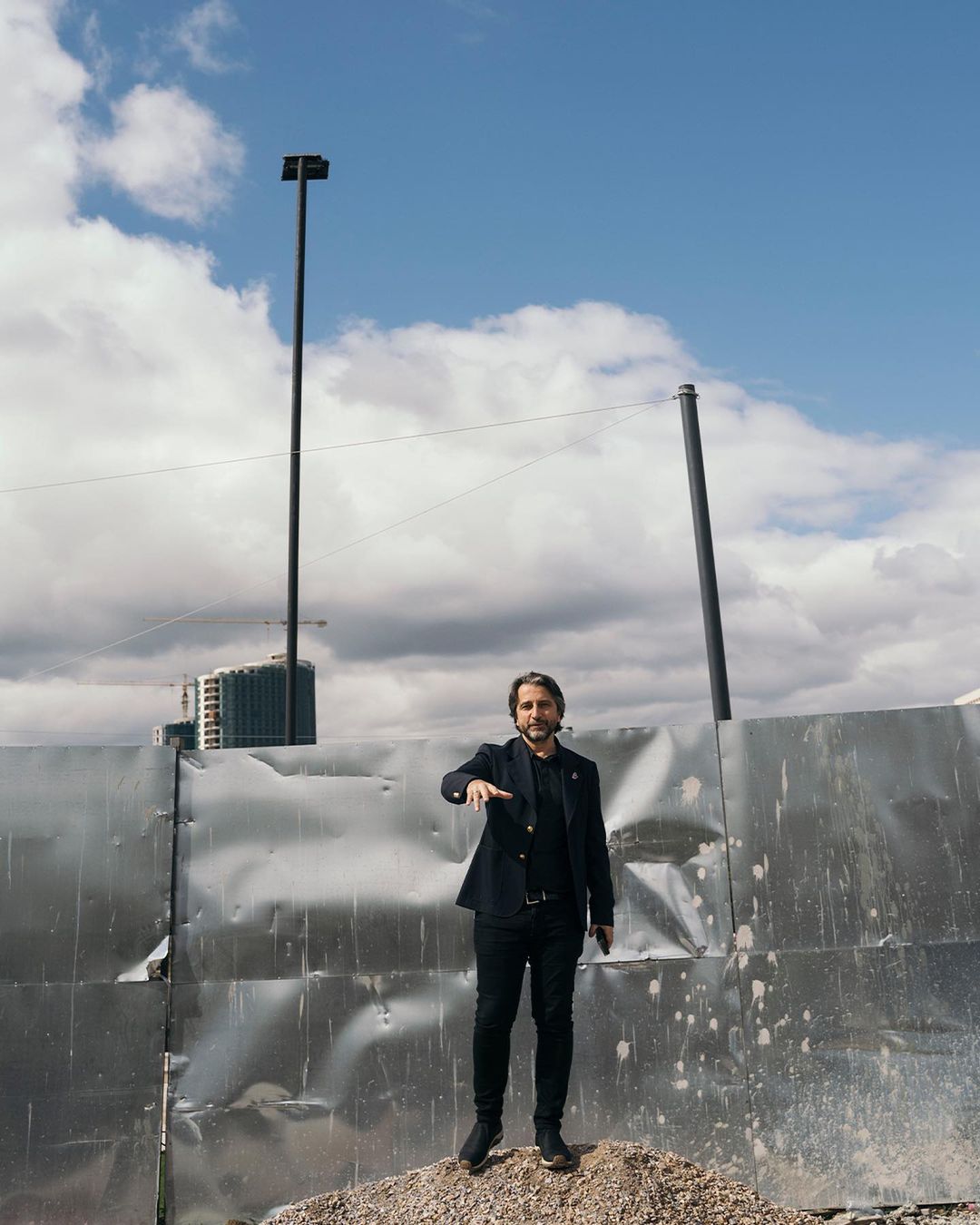Adult male in a black blazer stands on a gravel pile in front of a corrugated metal wall under a partly cloudy sky in an outdoor setting.