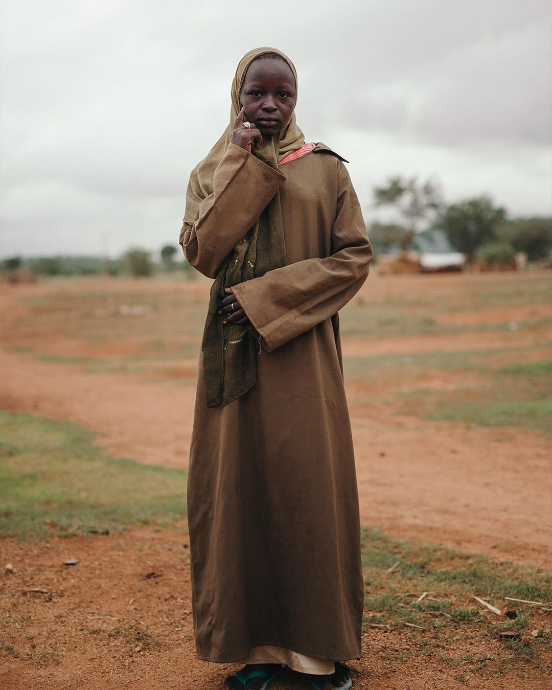 Full-length portrait of a dark-skinned adult female in a brown robe and headscarf, standing in a rural field with cloudy sky.