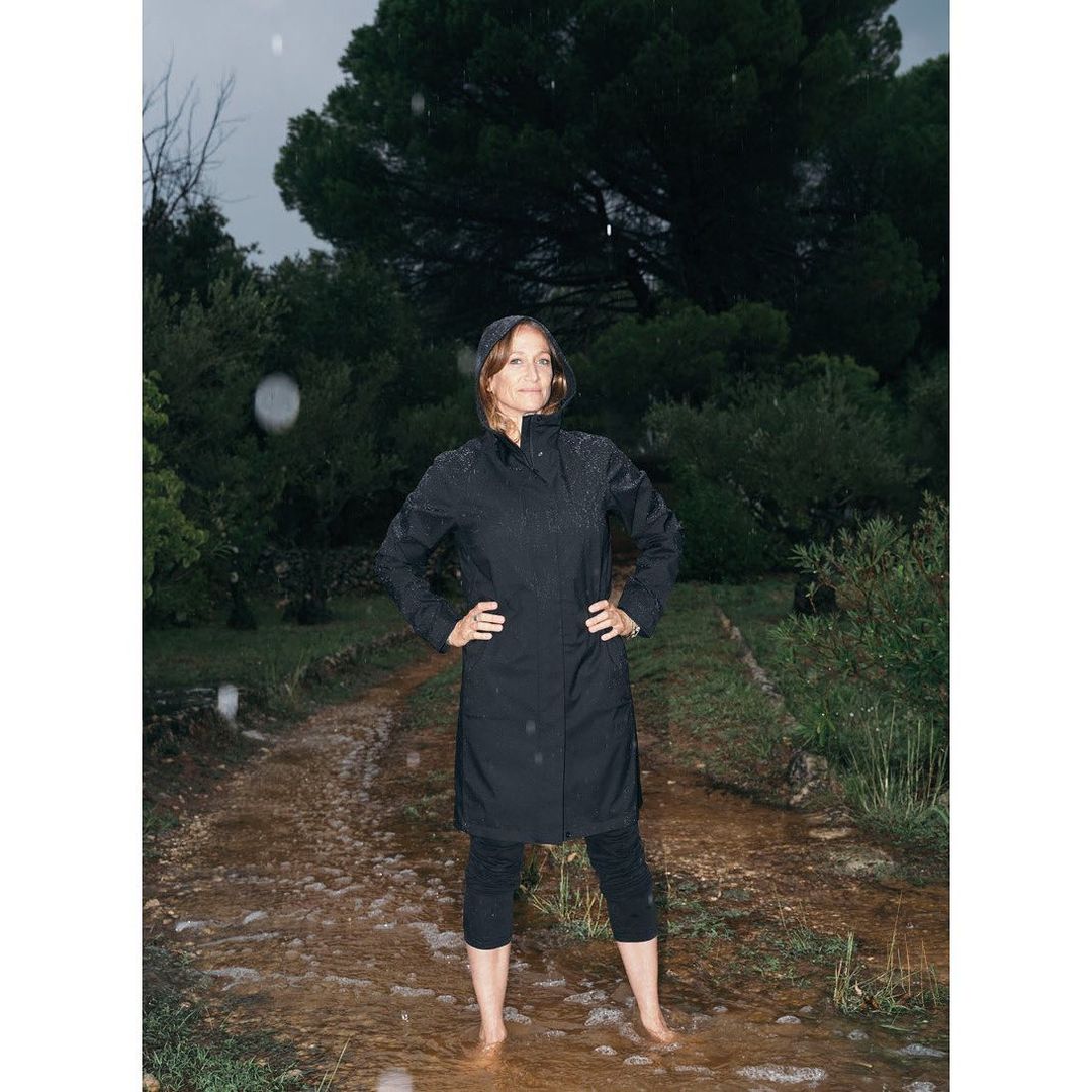 Woman in black raincoat standing barefoot in shallow water outdoors on a cloudy day, surrounded by green trees and foliage.