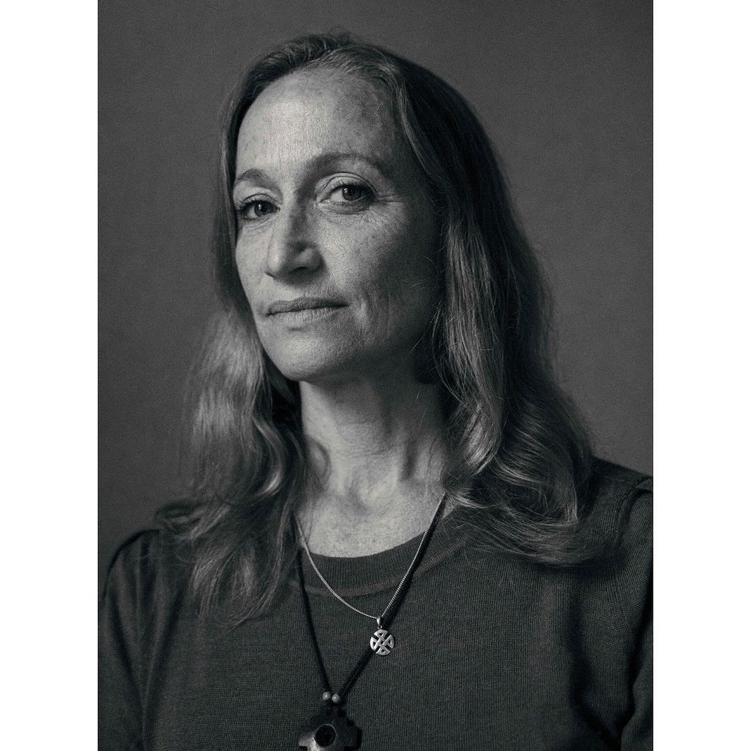 Monochrome portrait of an adult woman with long wavy hair, wearing a dark top and pendant necklaces against a plain background.