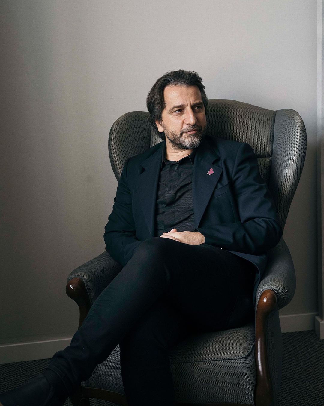 Adult male in a black suit relaxes in a gray armchair against a gray wall in an indoor environment with natural diffused lighting.