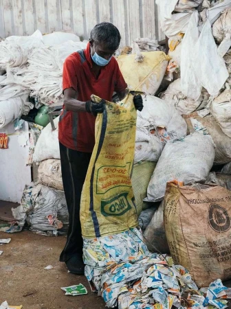 Man wearing a face mask sorts through plastic waste inside of a bag in a recycling facility, with bags of waste visible in the background.