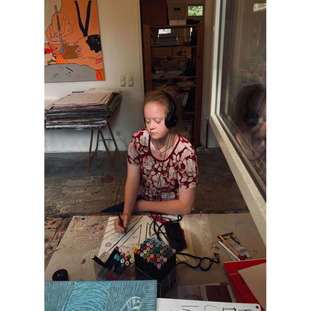 Young woman with headphones drawing at a table indoors surrounded by art supplies, featuring a painting and neatly stacked papers.