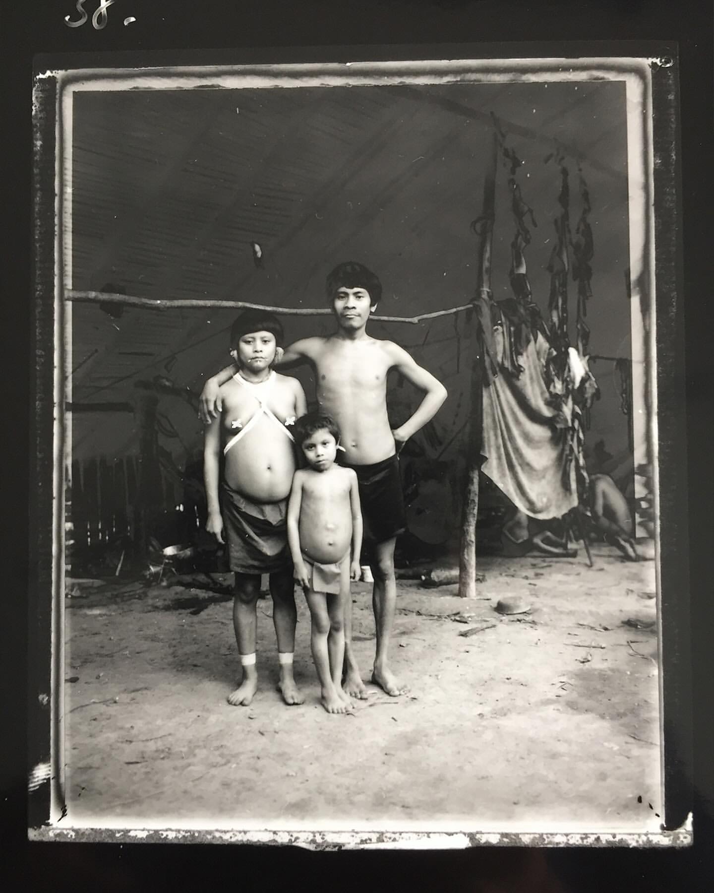 Black and white photo of three indigenous children standing outdoors in front of a tent-like structure, showcasing cultural elements.
