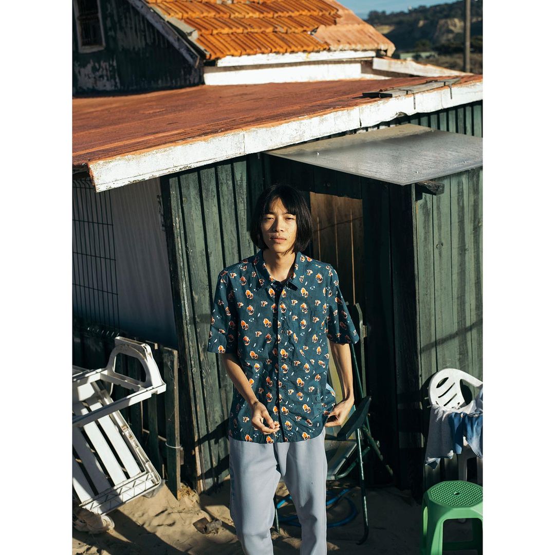A young man in a patterned shirt poses outdoors near a weathered building on a sunny day, showcasing casual style and a rustic backdrop.