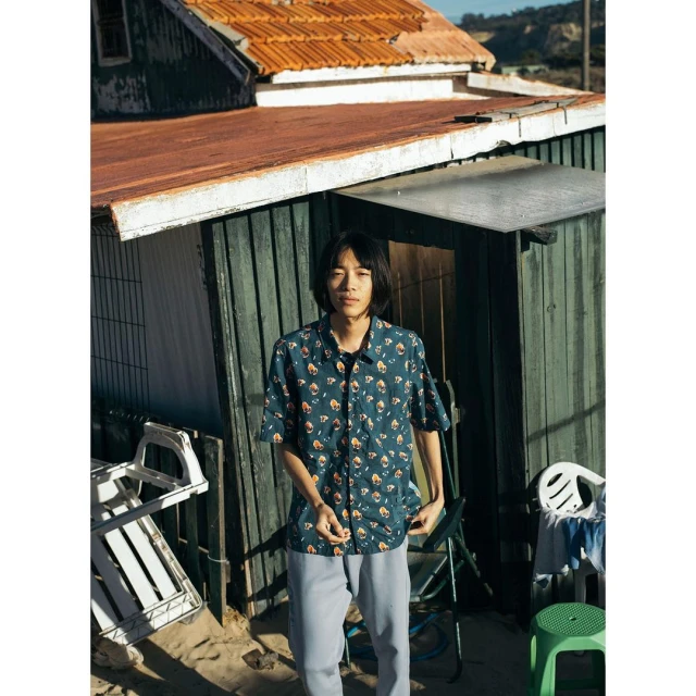A young man in a patterned shirt poses outdoors near a weathered building on a sunny day, showcasing casual style and a rustic backdrop.